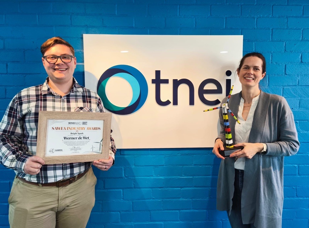 TNEI colleagues standing for a photo with their individual awards. On the left is Werner de Wet (Technical Consultant) with his finalist certificate for the Bright Spark Award. On the right is Zoë Lincoln (Principal Consultant) with her Watt-a-Woman Leadership Award. Between them is a sign with TNEI's logo on it.